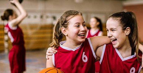 Zwei Basketball- Trickots tragende Mädchen lachen Arm in Arm und mit einem Basketball in der Hand in einer Turnhalle.| Sparkasse Hannover