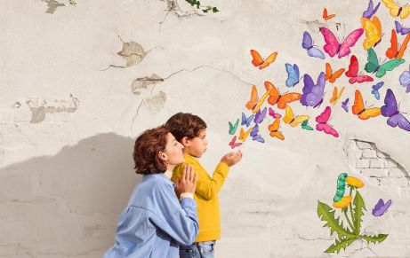 Frau kniet bei einem Kind. Beide stehen vor einer Wand. Außer der offenen Hand des Kindes steigt ein Schwarm Schmetterlinge auf | Sparkasse Hannover