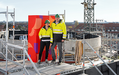 Ein Mann und eine Frau stehen auf einer Baustelle vor einer roten Stele | Sparkasse Hannover