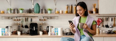 Junge Frau sitzt mit Smartphone und Tasse auf einer Kochinsel | Sparkasse Hannover